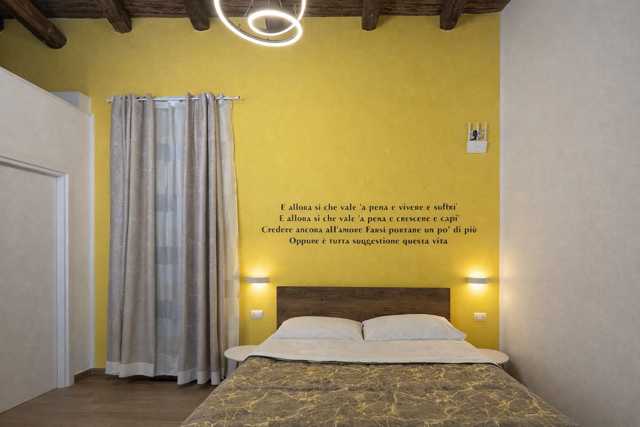 Camera con parete gialla e, sopra il letto, i versi di Allora sì
